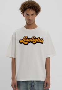 Νέος άντρας με σγουρά μαλλιά, φορώντας oversized λευκό t-shirt με κίτρινο και μαύρο κείμενο "Lowlights" και μπλε τζιν, στέκεται μπροστά σε απλό φόντο.