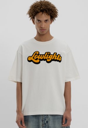 Junger Mann mit lockigem Haar, der ein übergroßes weißes T-Shirt mit gelbem und schwarzem "Lowlights"-Text und blauen Jeans trägt, steht vor einem schlichten Hintergrund.