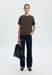 T-shirt di cotone marrone, vestibilità rilassata, maniche corte; abbinato a jeans svasati blu scuro. Tiene una borsa a tracolla in pelle nera con un motivo testurizzato.