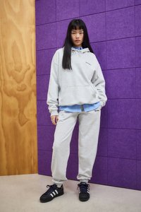 Donna in piedi contro un muro viola e di legno, indossando una felpa grigia chiara e pantaloni della tuta con scarpe nere e una maglietta blu sotto.