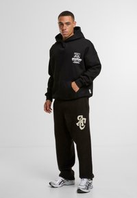 Sudadera negra con logo gráfico, bolsillo canguro en la parte delantera; combinada con pantalones de forro polar marrón oscuro con iniciales crema bordadas. Zapatillas deportivas blancas.