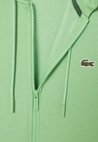Hoodie zippé vert clair en tissu doux, doté d'une fermeture éclair lisse et d'un petit logo de crocodile brodé sur la poitrine.