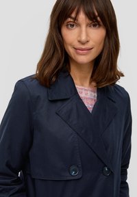 Dunkelblauer Trenchcoat mit doppelter Knopfreihe, großem Reverskragen und Knopfdetails. Tragen über einem hellen Pastellpullover.