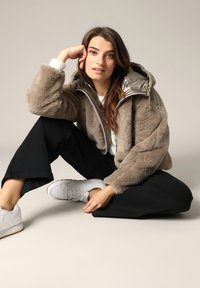 Veste cropped en fausse fourrure taupe, avec une doublure argentée et une capuche. Un pantalon noir à jambes larges et des baskets blanches complètent la tenue.