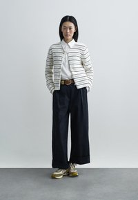 Gebreid vest met horizontale strepen, wit en zwart; wit overhemd met kraag, donkere broek met wijd uitlopende pijpen, en beige sneakers met groene accenten.