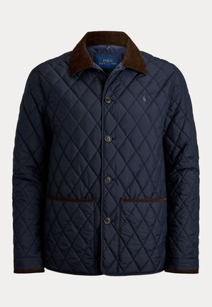 Hombre con gafas de sol, chaqueta acolchada azul marino, suéter de punto trenzado color oliva, vaqueros azul claro, botas de ante marrones, de pie con una mano en el bolsillo.