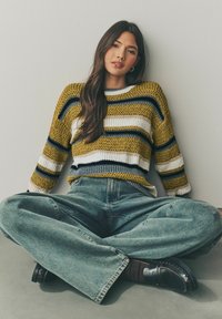 Strickpullover mit senfgelben, blauen, schwarzen und weißen Streifen, kombiniert mit hellblauen weiten Jeans und schwarzen Slippern, sitzt auf einem Boden.