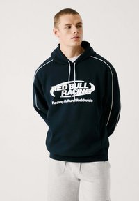 Sudadera con capucha de color marino con el logo en blanco de "RED BULL RACING" y acentos en blanco. Cuenta con capucha ajustable y textura suave, combinada con pantalones cortos grises.