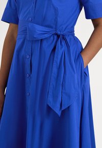 Ženska v modri obleki z gumbi, kratkimi rokavi in pasom, ki je privezan okoli pasu, z roko sproščeno v desnem žepu.