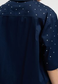 Camicia blu navy con maniche corte, decorata con piccole borchie quadrate argentate sparse sulla parte superiore della schiena e sulle maniche.