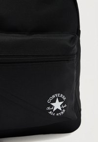 Μαύρο σακίδιο φτιαγμένο από ανθεκτικό ύφασμα, διαθέτει μπροστινή τσέπη με φερμουάρ και λευκό λογότυπο "Converse Chuck Taylor All Star" με σχεδιασμό αστεριού.