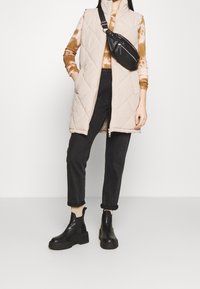 Gilet beige trapuntato con colletto alto, abbinato a un top marrone tie-dye e jeans neri. Stivaletti neri alla caviglia e piccola borsa a tracolla nera.