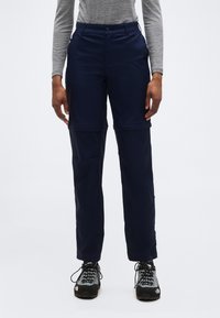 Άτομο που φοράει μπλε σκούρο παντελόνι μετατροπής με αποσπώμενα σορτς και γκρι παπούτσια πεζοπορίας, στέκεται μπροστά σε απλό φωτεινό φόντο.