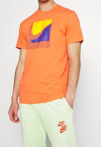 Muž v oranžovém tričku Nike s velkým žlutým a modrým logem a světlých zelených kalhotách se třemi oranžovými logy Nike.