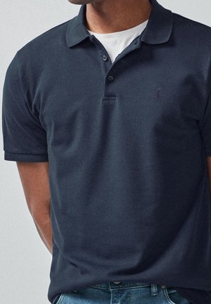 Mężczyzna w granatowej koszulce polo z rozpiętymi dwoma guzikami na białej koszulce pod spodem, w połączeniu z niebieskimi dżinsami.