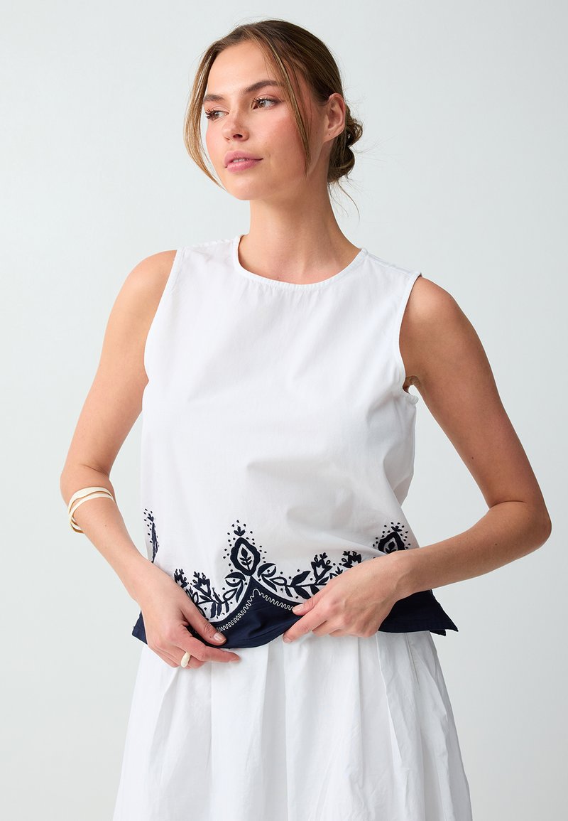 Femme portant un haut blanc sans manches avec une broderie florale bleu marine à l’ourlet, regardant sur le côté, cheveux attachés, fond clair.
