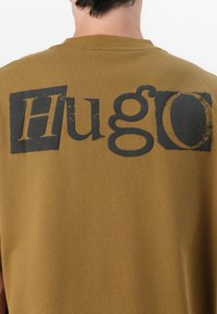 Bruine katoenen t-shirt met korte mouwen, voorzien van grote, geprinte zwarte tekst "Hu" en "go" op de achterkant. De tekst heeft een versleten effect.