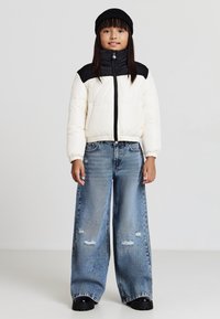 Steppjacke in Creme mit schwarzen Akzenten, hoher Kragen, Reißverschluss. Weit geschnittene, ausgefranste Jeans und schwarze Mütze vervollständigen das Outfit.