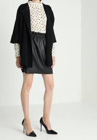 Manteau noir surdimensionné superposé à une blouse blanche à pois noirs, associé à une mini-jupe en cuir noir et des escarpins noirs à talons aiguilles.