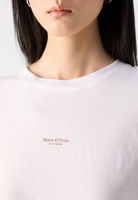 Balta medvilninė marškinėliai su apvali kaklo iškirpte, ant priekio esantis mažas rudas logotipas: "Marc O'Polo EST. IN STOCKHOLM".