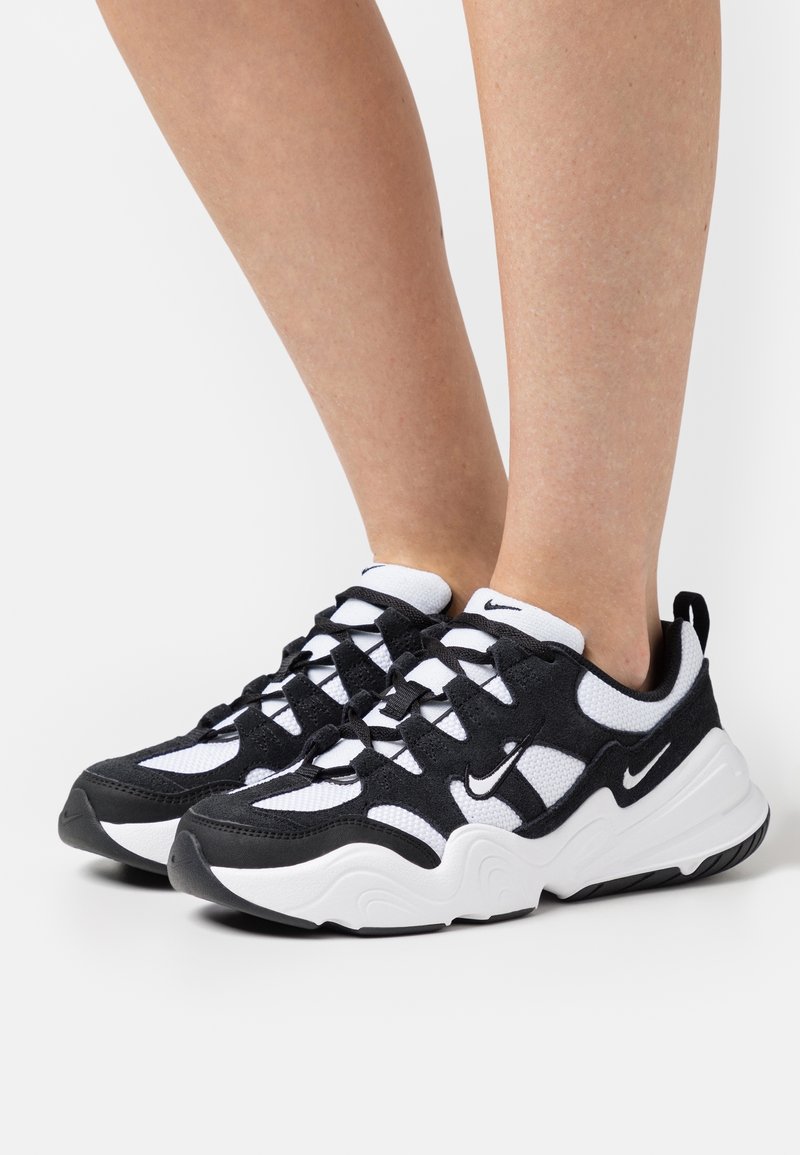 Jambes d'adulte portant des baskets Nike noires et blanches avec semelles texturées et design à lacets, debout sur une surface blanche unie.
