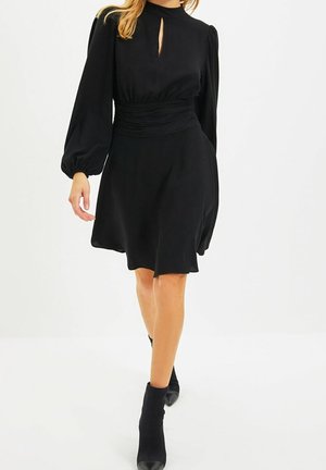 Vrouw die loopt, gekleed in een zwarte jurk met lange mouwen, een keyhole-halslijn en een geplooide taille, gecombineerd met zwarte enkellaarzen.