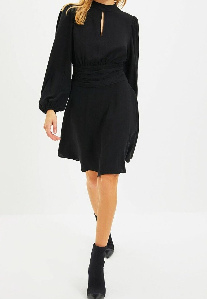 Femme marchant portant une robe noire à manches longues avec un col en forme de goutte et une taille froncée, associée à des bottines noires.