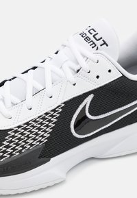 Sorte og hvite sneakers med mesh-overdel, teksturert design, fremtredende merkevare, og en slank form med gummisåle.