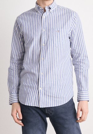 Hombre vistiendo una camisa de manga larga con rayas verticales en azul claro y blanco, con botones, y jeans azul oscuro, de pie contra un fondo liso.