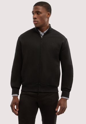Mann trägt eine schwarze Zip-up-Jacke über einem gestreiften Hemd, steht vor einem einfarbigen hellen Hintergrund und schaut nach links.