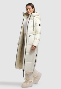 Lang, puffet hvid frakke med høj krave, sorte lynlåse og sidelommer. Kombination med lyse, isolerede støvler. Glat tekstur.