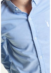 Personne portant une chemise bleu clair boutonnée avec un petit logo de cerf blanc sur la poitrine, visible du cou à la taille.