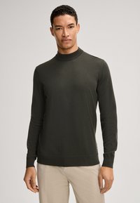 Dunkelgrüner, langärmeliger Rollkragenpullover aus glattem Strickmaterial, mit gerippten Bündchen und Saum. Über hellen Hosen getragen.