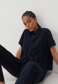 Camicia blu navy a maniche corte con bottoni, realizzata in un tessuto testurizzato, abbinata a pantaloni coordinati. La camicia presenta un colletto e maniche arrotolate.