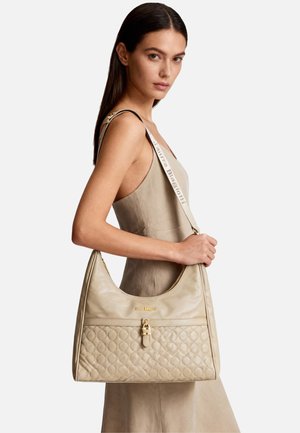 Donna in abito beige che porta una grande borsa a tracolla beige trapuntata con chiusura dorata e tracolla con logo sulla spalla.