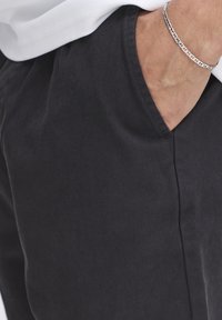 Black cotton pants with a smooth texture, featuring side pockets and a relaxed fit. A silver chain bracelet is visible on the hand.
