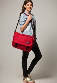 Bolsa de mensajero de lona roja con cierre de solapa, correa ajustable y un bolsillo con cremallera en el frente. Acompañada por un modelo que lleva una chaqueta de mezclilla clara.
