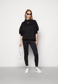 Juodas didelio dydžio sportinis džempers su baltu Nike logotipu, derinamas su juodomis legginsais ir baltomis sportinėmis bateliais. Aksesuarai apima mažą baltą rankinę ir saulės akinius.