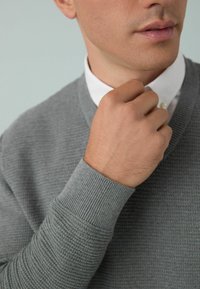 Suéter gris con textura y puños acanalados, llevado sobre una camisa blanca con cuello. Primer plano de una mano ajustando el cuello.