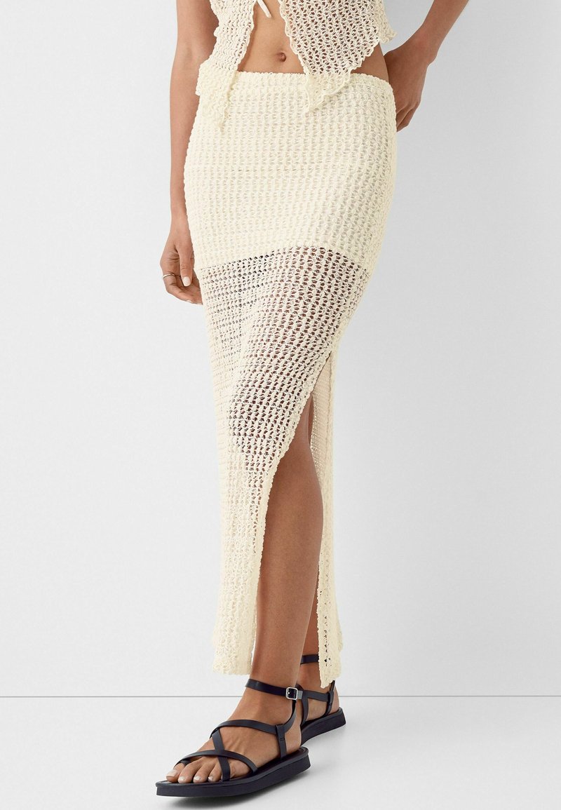 Femme portant une jupe midi en crochet crème avec une fente latérale, associée à des sandales plates noires à lanières, debout devant un fond uni.