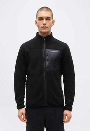 Homme portant une veste polaire noire zippée avec une poche poitrine zippée et le logo "Peak Performance" sur la poche sur un fond uni.
