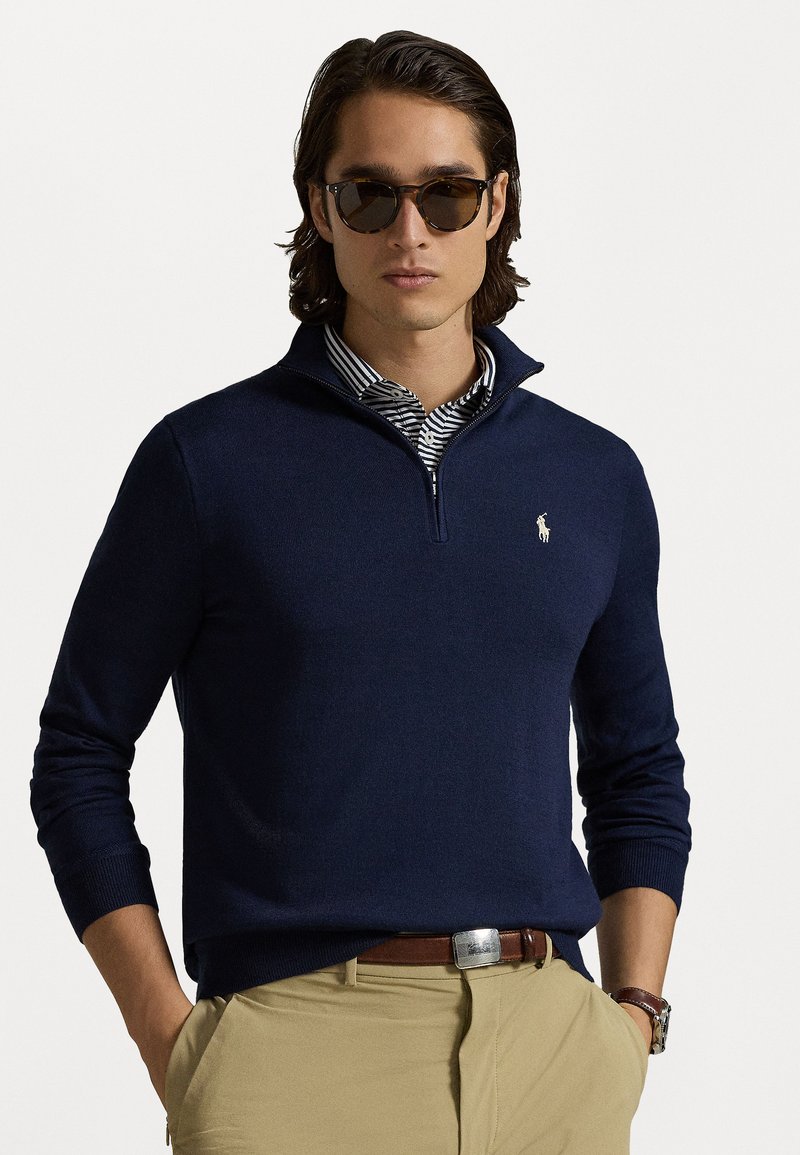 Pulover din tricot cu guler tip zip, de culoare bleumarin, realizat dintr-un material neted, având un mic logo brodat, purtat peste o cămașă cu guler în dungi.