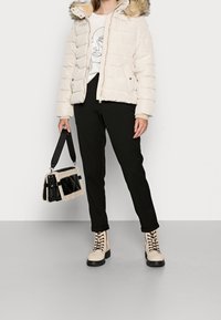 Kvinna i en krämfärgad pufferjacka med pälsmudd, vit t-shirt med grafik, svarta byxor, krämfärgade snörboots, som håller en svart och krämfärgad handv äska.