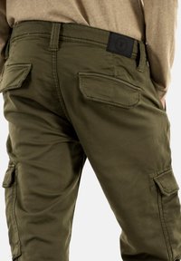 Olijfgroene cargobroek heeft een relaxte pasvorm, meerdere zijzakken en een zwart logo-opdruk op de tailleband. Gemaakt van duurzaam materiaal met een gladde textuur.