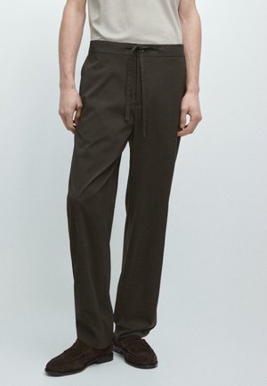 Homme portant un pantalon à cordon de serrage marron foncé et des chaussures marron foncé, debout devant un fond clair uni.