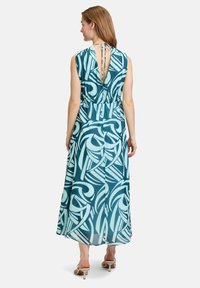 Maxi-jurk in teal met abstracte lichtblauwe patronen. Heeft korte mouwen, een striksluiting aan de achterkant en een luchtige, semi-doorzichtige stof.