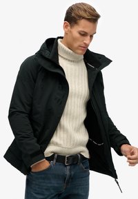 Schwarze wasserdichte Jacke mit hohem Kragen, Reißverschlusstaschen und "SUPERDRY"-Markierung; getragen über einem cremefarbenen gerippten Pullover und blauen Jeans.