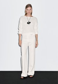 Witte gebreide trui met ribbelzoom, gecombineerd met witte wijde broeken en zwarte open sandalen. Zwarte hangende ketting inbegrepen.