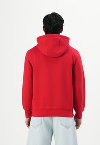 Uomo con capelli scuri che indossa un hoodie rosso e jeans azzurri, in piedi di spalle contro uno sfondo bianco semplice.