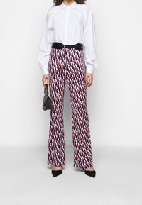Pantalons évasés à motifs en noir, blanc et rose, présentant des formes géométriques. Associés à une chemise blanche à boutons et une ceinture noire.